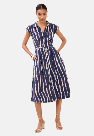 Naine kandmas varrukateta mereväe- ja valgekirju midi-kleiti vöögaiaga, seisab käed taskutes, jalas beežid rihmikujalatsid.