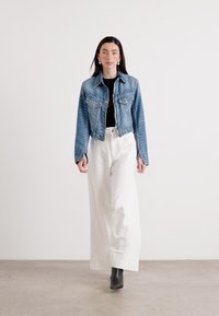 Mudel kõnnib enesekindlalt denim-jakiga, musta särgi ja valgete laia säärega pükstega, minimalistlikus taustas ja stiilsete ankle bootsidega.