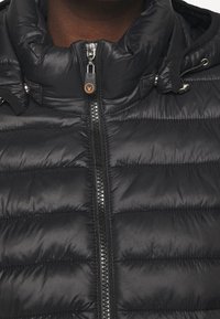 Czarna pikowana kurtka typu puffer z wysokim kołnierzem, wyraźnym poziomym przeszyciem oraz przednim zamkiem błyskawicznym z metalowym suwakiem i logo.