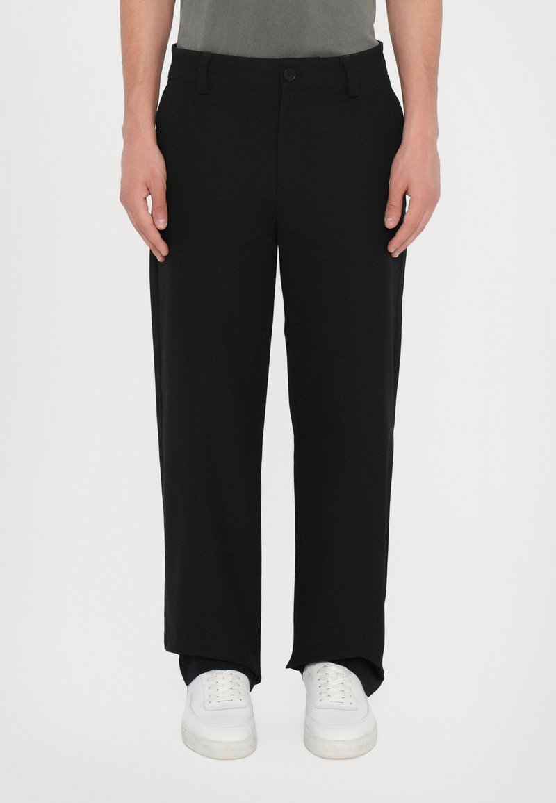 Personne portant un pantalon noir droit et des baskets blanches, debout sur un fond clair uni.