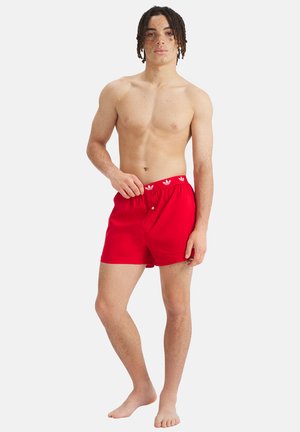 Junger Mann mit mittlerer Hautfarbe und Dreadlocks, der rote Adidas-Boxershorts trägt und barfuß vor einem schlichten weißen Hintergrund steht.
