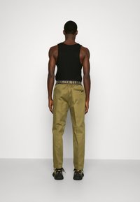 Man facing away wearing black tank top, olive-green trousers with branded waistband, and black sneakers with gold accents, standing on gray floor.