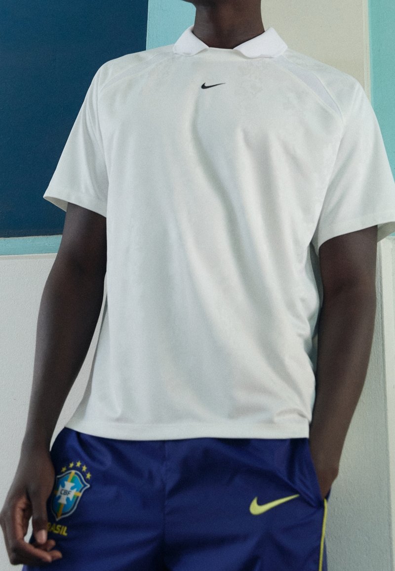 Polo shirt Nike blanc avec un col structuré, orné d'un petit logo sur la poitrine. Porté avec un short bleu avec des détails jaunes.