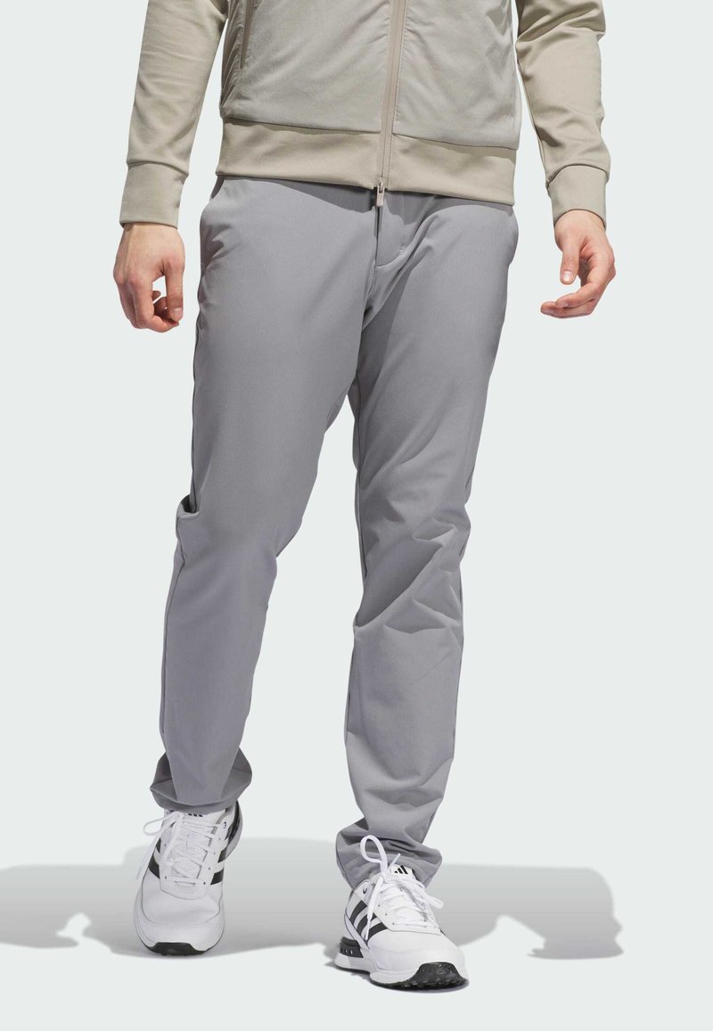 Pantalons de sport gris clair avec une coupe fuselée, texture lisse, poches latérales et taille élastique, associés à des baskets blanches.
