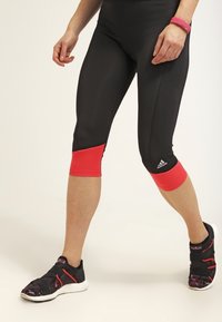 Person iført sorte og røde Adidas capri leggings og sorte løbesko med hvide såler, stående på en lys baggrund.
