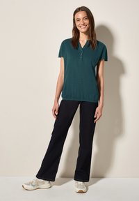 Polo-shirt teal à manches courtes avec une patte de boutonnage à trois boutons, coupe décontractée et ourlet élastique, associé à un jean foncé et des baskets blanches.