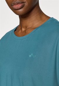 Τιρκουάζ κοντομάνικη μπλούζα με στρογγυλή λαιμόκοψη και διακριτικό πουα σχέδιο στο αριστερό μέρος, κατασκευασμένη από μαλακό, ελ αφρύ ύφασμα.