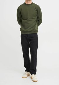 Olijfgroene sweatshirt met ronde hals, lange mouwen en ribbels bij de manchetten, gecombineerd met zwarte broeken en witte schoenen met tan accenten.