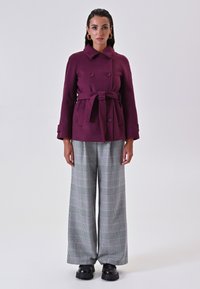 Cappotto bordeaux doppiopetto con vita legata, abbinato a pantaloni grigi a quadri con gamba ampia e scarpe nere chunky.