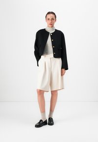 Černá zkrácená bunda se zlatými knoflíky, pruhovaný rolák, smetanové plisované šortky a černé mokasíny na béžových ponožkách. Minimalistický design.