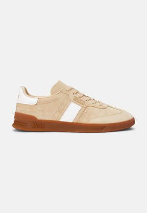 Beige suede sneakers with white stripes, rubber sole, and tan accents. Features a textured body and a logo on the side.