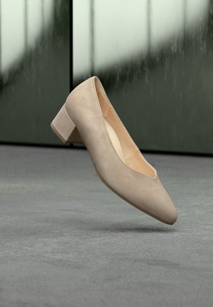 Zapato beige de ante con tacón bajo y punta afilada, flotando sobre un suelo gris con fondo borroso de pared verde reflectante.