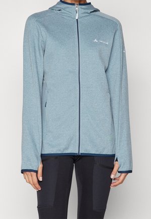 Veste à capuche bleu clair avec fermeture éclair, avec trous pour les pouces, portée avec un pantalon noir, logo à l'avant sur la poitrine et la manche.