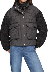 Vrouw in een zwarte gewatteerde jas met drukknoopsluiting, met textuur op de mouwen, en een blauwe spijkerbroek, staande met haar handen in de zakken.