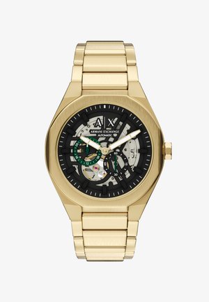 Montre-bracelet en métal doré avec cadran squelette noir, aiguilles blanches, sous-cadrans verts et marquage Armani Exchange, mouvement automatique visible.