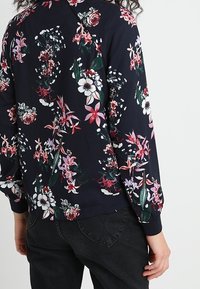 Marineblauw long-sleeve shirt met een veelkleurige bloemenprint, gedragen met zwarte jeans, achteraanzicht van een persoon die tegen een effen achtergrond staat.