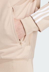 Light beige zippered pocket on a textured fabric, integrated into a long-sleeve top with three white stripes along the sleeve.