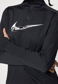 Sort quarter-zip langærmet top lavet af glat, strækbart materiale, med et hvidt Nike-logo på brystet og tommelhuller i manchetterne.