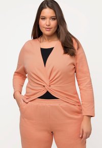 Top a maglia color pesco con maniche lunghe e dettaglio a nodo attorcigliato in vita, abbinato a pantaloni pesco coordinati. Canottiera nera visibile sotto.