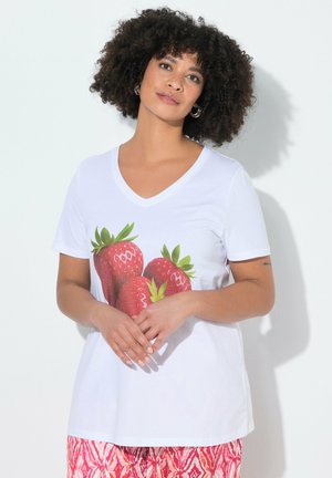 Frau mit lockigem Haar, die ein weißes V-Ausschnitt-T-Shirt mit großem Erdbeerdruck und rote gemusterte Hose trägt, steht vor einer weißen Wand.