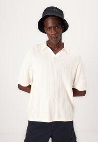 Polo en tricot beige clair à manches courtes, avec un col en V et un motif de côtes texturé. Associé à un short cargo bleu marine foncé et un chapeau de seau noir.