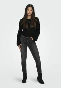 Maglione nero a maglia con maniche lunghe, abbinato a jeans slim-fit grigi scuri e stivali neri alla caviglia. Modello in posa su uno sfondo semplice.