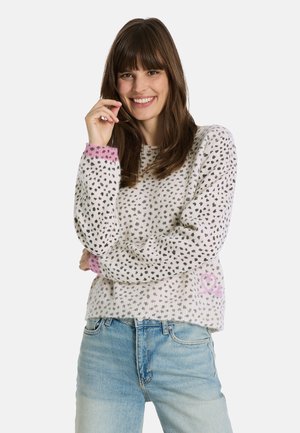 Strickpullover in Weiß mit schwarzen Punkten, pinken Bündchen und einem pinken Herzdetail. Lange Ärmel und lockerer Schnitt. Kombiniert mit hellblauen Jeans.