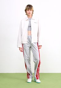 Balta strīpota jaka, pelēks īss tops, pelēkas bikses ar sarkaniem sānu svītrām un logo, baltas kedas ar tīklu akcentiem, uz zaļa grīdas.