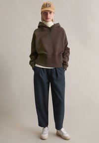 Braunes Kapuzen-Sweatshirt, beiger Cap mit "15", dunkelblaue Hose und weiße Turnschuhe. Glatter Stoff, lockerer Schnitt und minimalistisches Design.