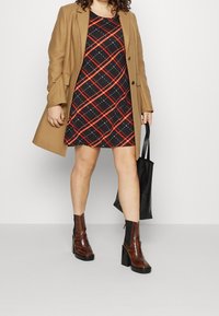 Cappotto marrone sopra un vestito nero con motivo a quadri rossi; stivali con tacco marroni; tiene una borsa tote nera. Sfondo minimalista.