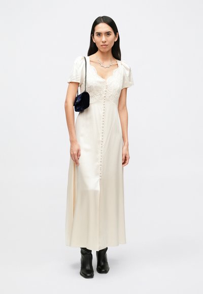 Vestido maxi em cetim creme com detalhes em renda, mangas curtas bufantes, abotoamento central e uma fenda. Usado com uma pequena mala azul-marinho e botas pretas.