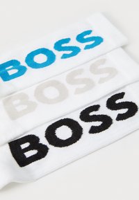 Drei Paar weißer Socken mit unterschiedlich farbigem "BOSS"-Text: hellblau, beige und schwarz, die eine gestrickte Textur und fette Buchstaben zeigen.