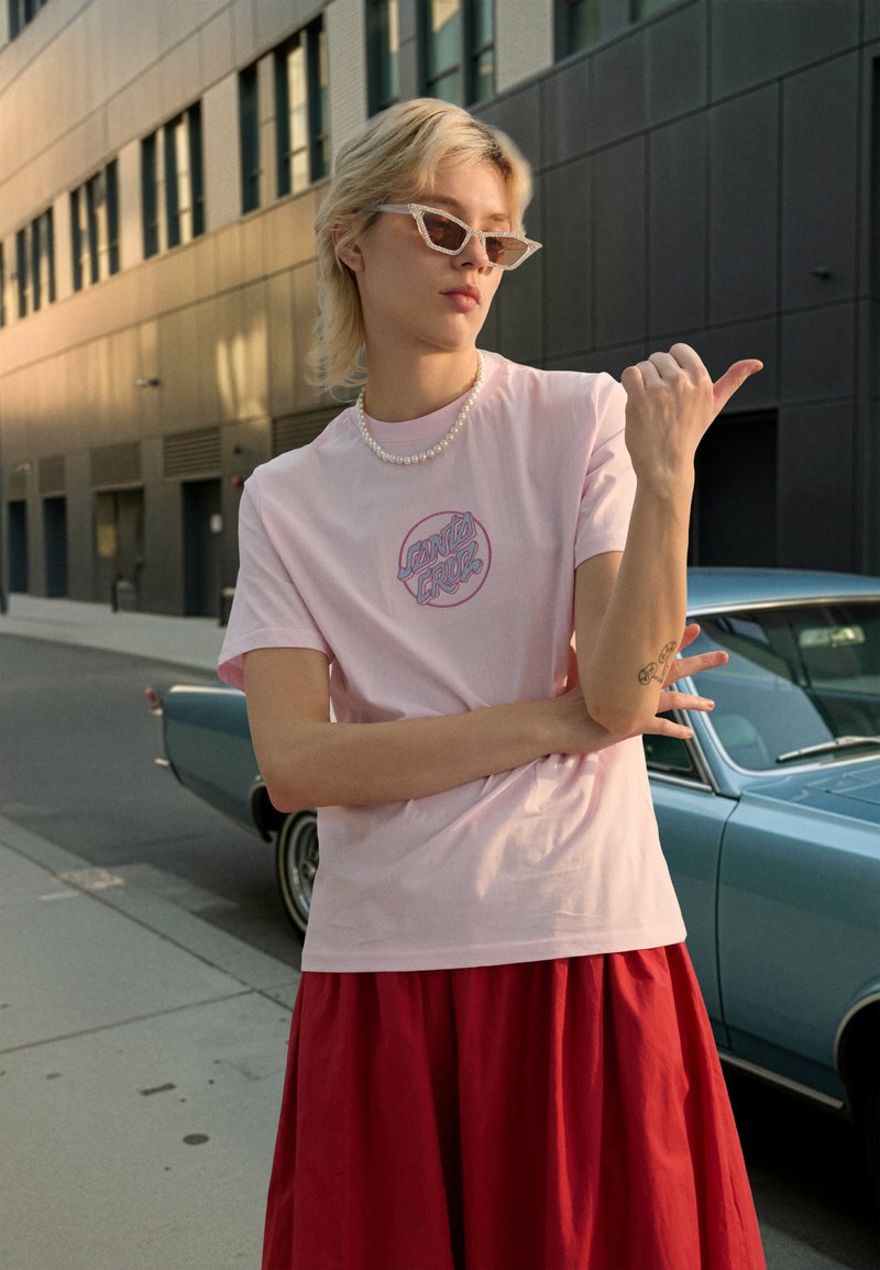 Ljusrosa bomulls-t-shirt med cirkulärt logga, bärs med röd kjol, pärlhalsband och kantiga solglasögon, mot en stadsmiljö.