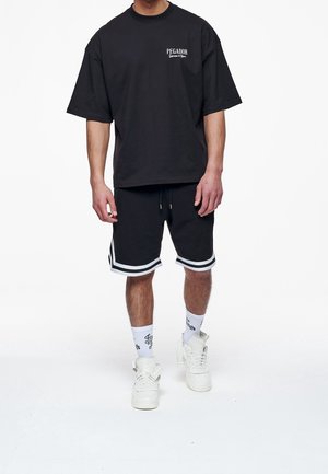Uomo che indossa una t-shirt nera oversize con la scritta "PEGADOR", pantaloncini neri con strisce bianche, calzini bianchi con scritta e sneaker bianche alte.