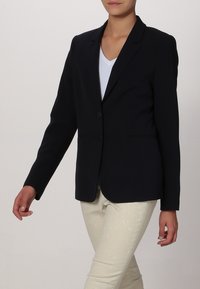Blazer bleu marine avec une fermeture à un bouton, des revers crantés, des poches latérales et une texture lisse, associé à un pantalon de couleur claire.