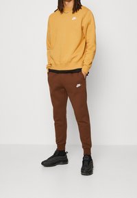 Keltainen collegepaita, jossa valkoinen Nike-logo, yhdistettynä ruskeisiin jogger-housuihin, joissa pieni valkoinen Nike-logo, ja mustat urheilukengät.