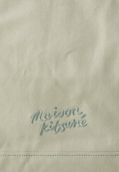Tela de lona verde claro con el texto bordado "maison kitsuné" en un tono complementario. Textura suave con un sutil detalle de costura.