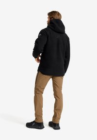 Svart fleece-hoodie med huva, texturerad yta, logotyp på ryggen, kombinerad med tanfärgade byxor och svarta träningsskor.