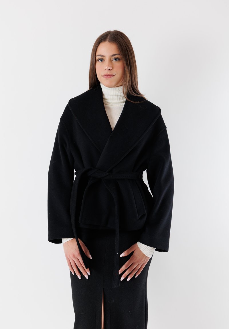 Cappotto nero avvolgente in un tessuto testurizzato, dotato di ampi rever, una cintura da annodare e maniche lunghe. Indossato sopra un dolcevita bianco, con una gonna sotto.