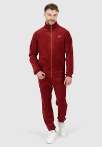 Roter Velours-Trainingsanzug mit Reißverschlussjacke, hohem Kragen und goldenen Logo. Eng anliegende Hosen mit Seitentaschen. Model trägt weiße Turnschuhe.