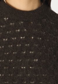 Pull tricoté marron foncé avec un motif texturé et de petits entrelacs ouverts. Col rond et matière douce et pelucheuse visible en gros plan.