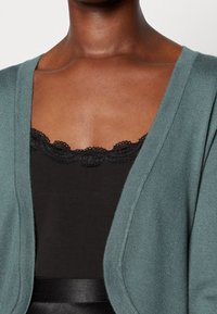Gråblå stickad cardigan med öppen framdesign, bärs över en svart topp med spetsdetalj längs halsringningen. Mjuk textur.