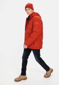 Rød polstret jakke med hætte, der har en lynlås og to sidelommer. Båret over en mørkeblå top og parret med mørkejeans og brune støvler.