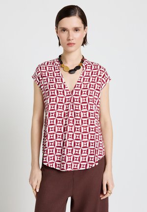 Femme portant un chemisier à manches courtes à motif géométrique rouge et blanc, un pantalon marron, et un collier épais noir et or, debout devant un fond blanc.