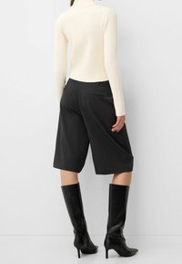 Nainen, jolla on päällään valkoinen ribbattu pitkähihais-toppi, mustat polvipesäiset leveät shortsit ja mustat korkeat polvisukat, seisoo tasaisella seinällä.