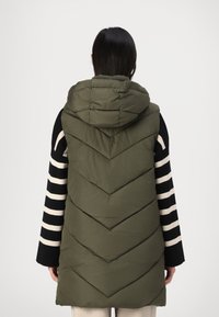 Gilet matelassé vert olive avec une capuche, présentant un design en chevrons. Le mannequin porte une chemise à rayures noires et blanches à manches longues en dessous.