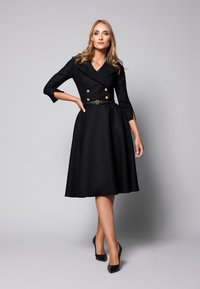 Czarna sukienka do kolan, dwurzędowa, z złotymi guzikami, rękawami do łokci, paskiem w talii i rozkloszowanym dołem; noszona z czarnymi szpilkami.