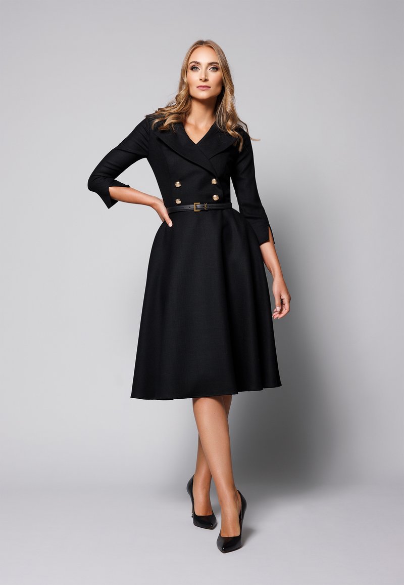 Czarna sukienka do kolan, dwurzędowa, z złotymi guzikami, rękawami do łokci, paskiem w talii i rozkloszowanym dołem; noszona z czarnymi szpilkami.