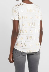 Osoba oblečená v bílé krátké košili s zlatým textem "TRUE RLGN" a šedých džínách, zobrazena zezadu.