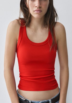 Jeune femme portant un débardeur rouge côtelé ajusté et un jean bleu clair avec une ceinture noire sur un fond clair uni.